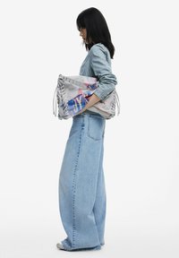 Große Stoffhandtasche in hellen Farben mit Schnürseiten, versehen mit bedruckten Designs in Blau und Pink. Das Model trägt ein Denim-Outfit.