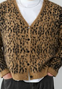 Bruine, fluffy cardigan met zwarte luipaardvlekken, een v-hals en zwarte knopen, gelaagd over een wit shirt en grijze broek.