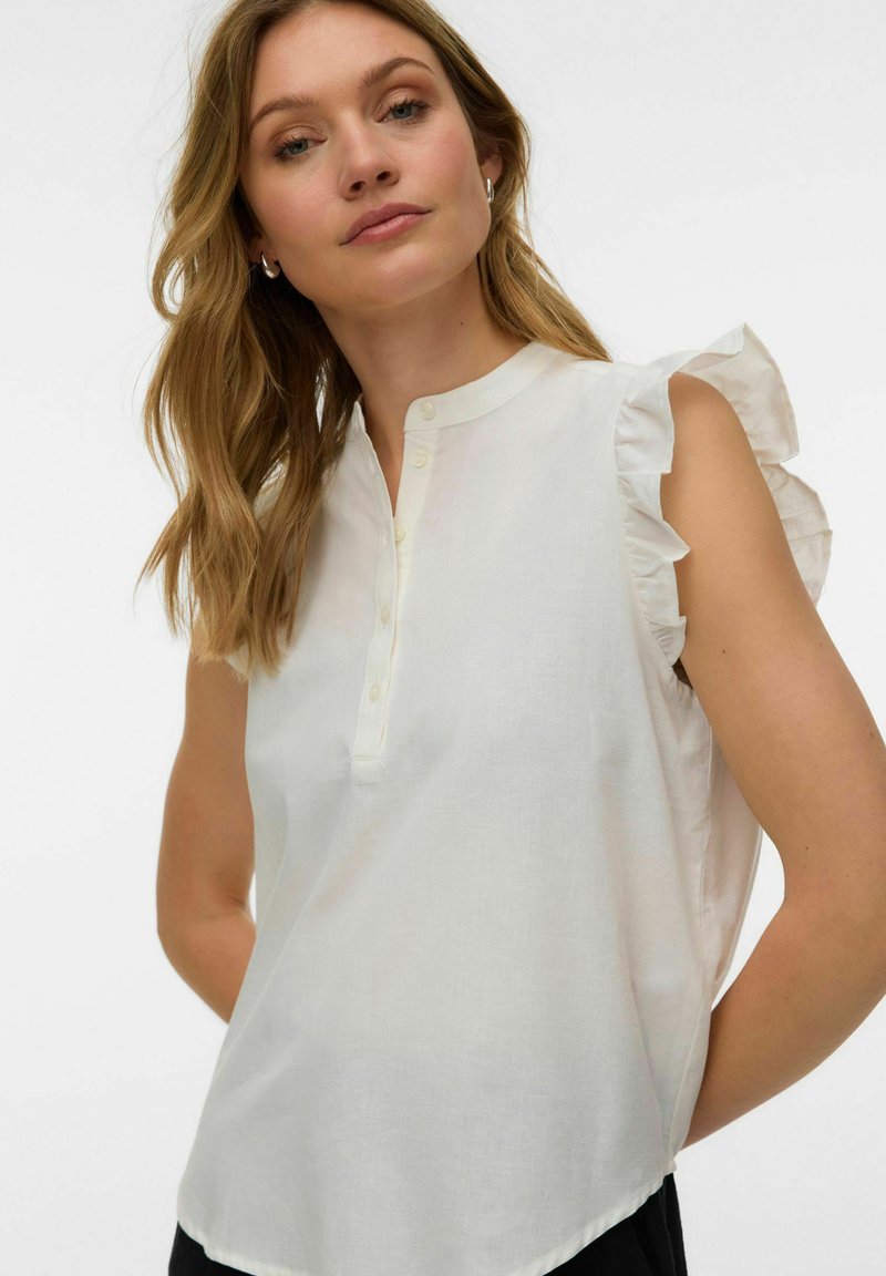 Femme aux cheveux blonds ondulés portant un chemisier blanc sans manches avec des épaules volantées et un col boutonné, debout devant un fond uni.
