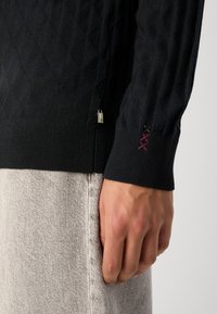 Schwarzer gestrickter Pullover mit einem Rautenmuster, gerippten Bündchen und einem kleinen metallischen Logo-Detail an der Seite. Hand nahe einem hellgrauen Stoff.