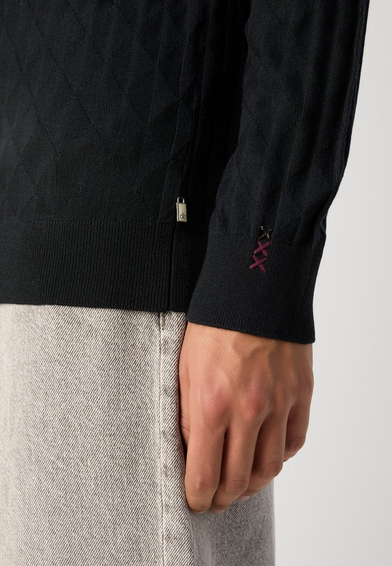 Zwarte gebreide trui met een ruitenpatroon, geribde manchetten en een klein metalen logo aan de zijkant. Hand dicht bij lichtgrijze stof.