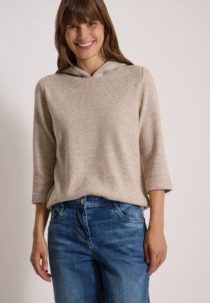 Femme souriante portant un pull à capuche beige à manches trois-quarts et un jean bleu sur un fond clair uni.