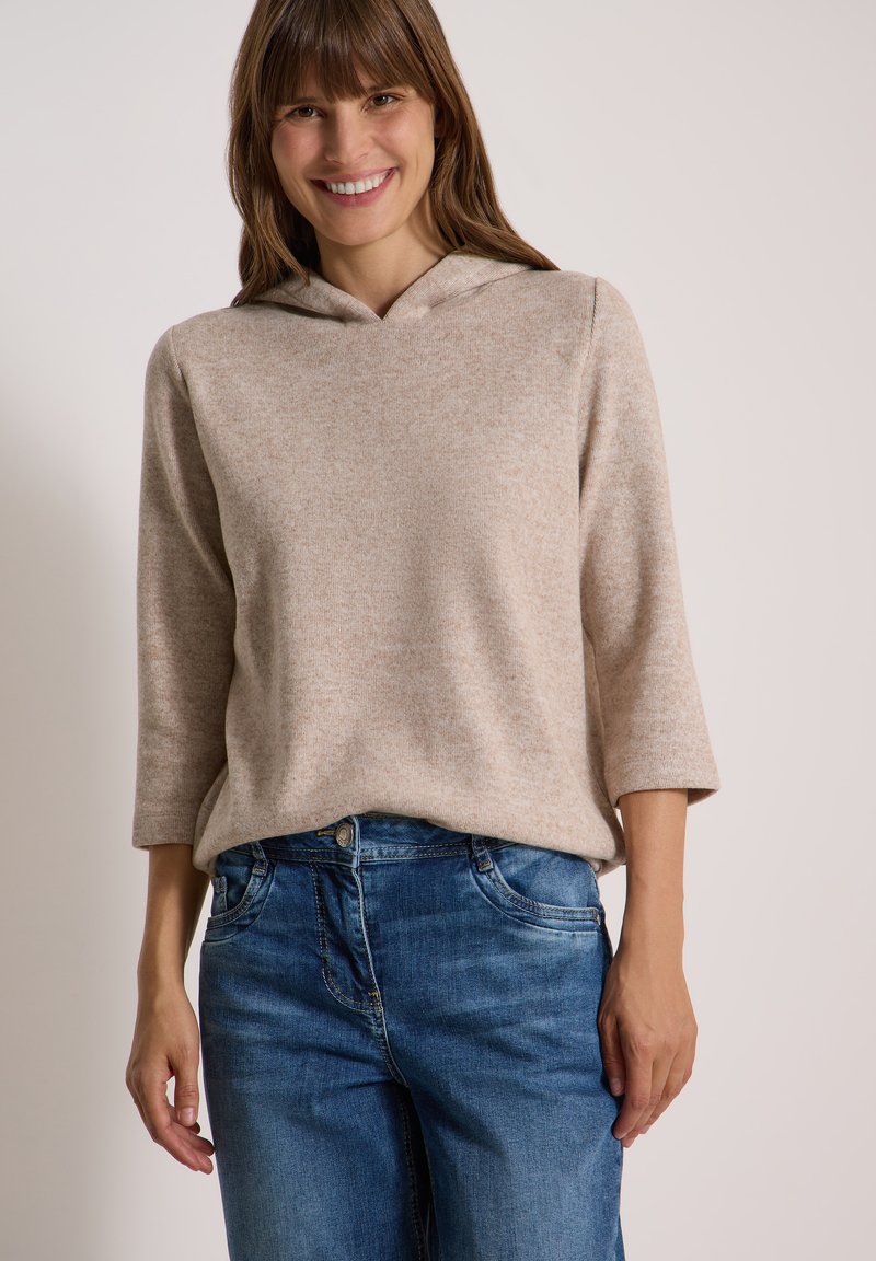 Femme souriante portant un pull à capuche beige à manches trois-quarts et un jean bleu sur un fond clair uni.
