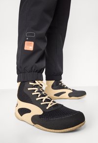 Black and beige athletic shoes with a mesh design, flat laces, textured soles, and a smooth finish. Black joggers with a logo patch.
