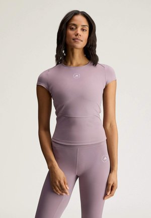 Donna che indossa una maglia sportiva aderente a maniche corte color malva e leggings con un logo discreto sul petto e sulla coscia, in piedi su uno sfondo neutro.