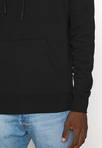 Felpa nera con cappuccio realizzata in un materiale morbido, dotata di tasca a marsupio e polsini a costine, abbinata a jeans in denim blu chiaro.
