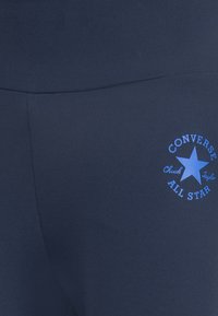 Leggings en tissu extensible bleu marine avec un logo Converse All Star arrondi en bleu sur le côté, présentant une texture lisse et un design ajusté.