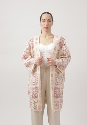 Vrouw staat en draagt een lange, open voorkant gehaakte cardigan met roze en crème kleuren, over beige broek en een wit shirt.