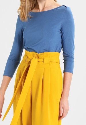 Kvinde iført en blå langærmet top stoppet ned i højtaljede gule bukser med et bredt bælte bundet i en sløjfe.
