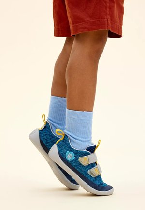 Blaue und graue Kinderschuhe mit strukturiertem Strickobermaterial, verstellbaren Klettverschlüssen und gelben Akzenten, kombiniert mit hellblauen gerippten Socken.