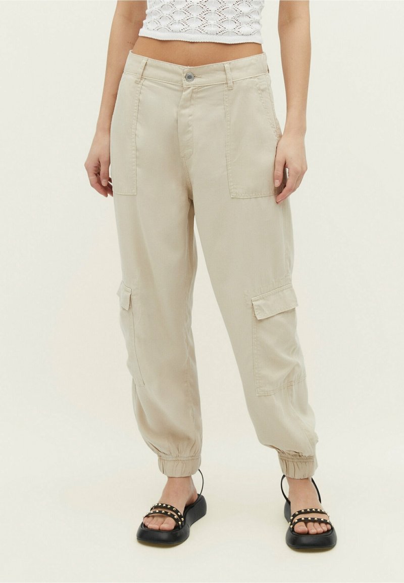 Femme portant un pantalon cargo beige avec des poignets élastiques, un top court en maille blanc et des sandales noires à brides cloutées.