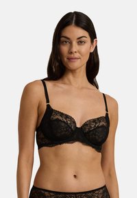 Černá krajková podprsenka s průsvitnými panely, s kosticemi, nastavitelnými ramínky, vlnkovanými okraji a detaily ve zlaté barvě.