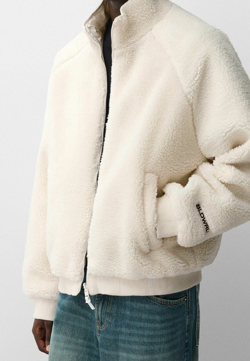 Chaqueta de forro polar en color blanco roto, con un alto cuello, cierre de cremallera, puños acanalados y un detalle de logo en la manga. Textura suave.