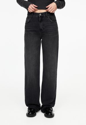 Personne portant un jean noir ample, un haut à manches longues noir et des chaussures noires brillantes, debout devant un fond blanc uni.
