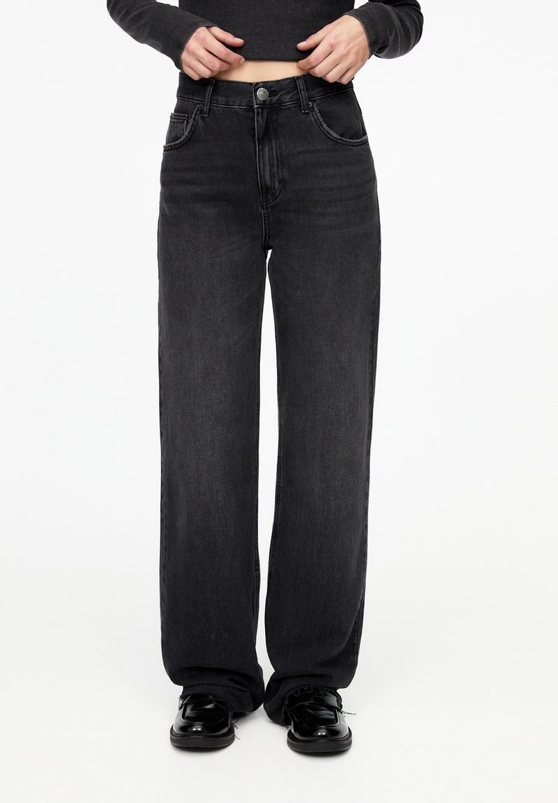 Personne portant un jean noir ample, un haut à manches longues noir et des chaussures noires brillantes, debout devant un fond blanc uni.