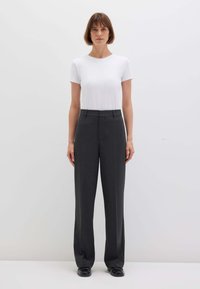 Donna con capelli corti castani che indossa una maglietta bianca semplice a maniche corte, pantaloni alti grigio scuro e scarpe nere, in piedi su uno sfondo bianco.
