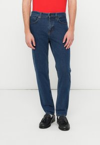 Tumed sinised teksapüksid sirge sääre disainiga, viie taskuga ja klassikalise vööümbrusega. Sobitatud mustade loafers-kingade ja punase toppiga.