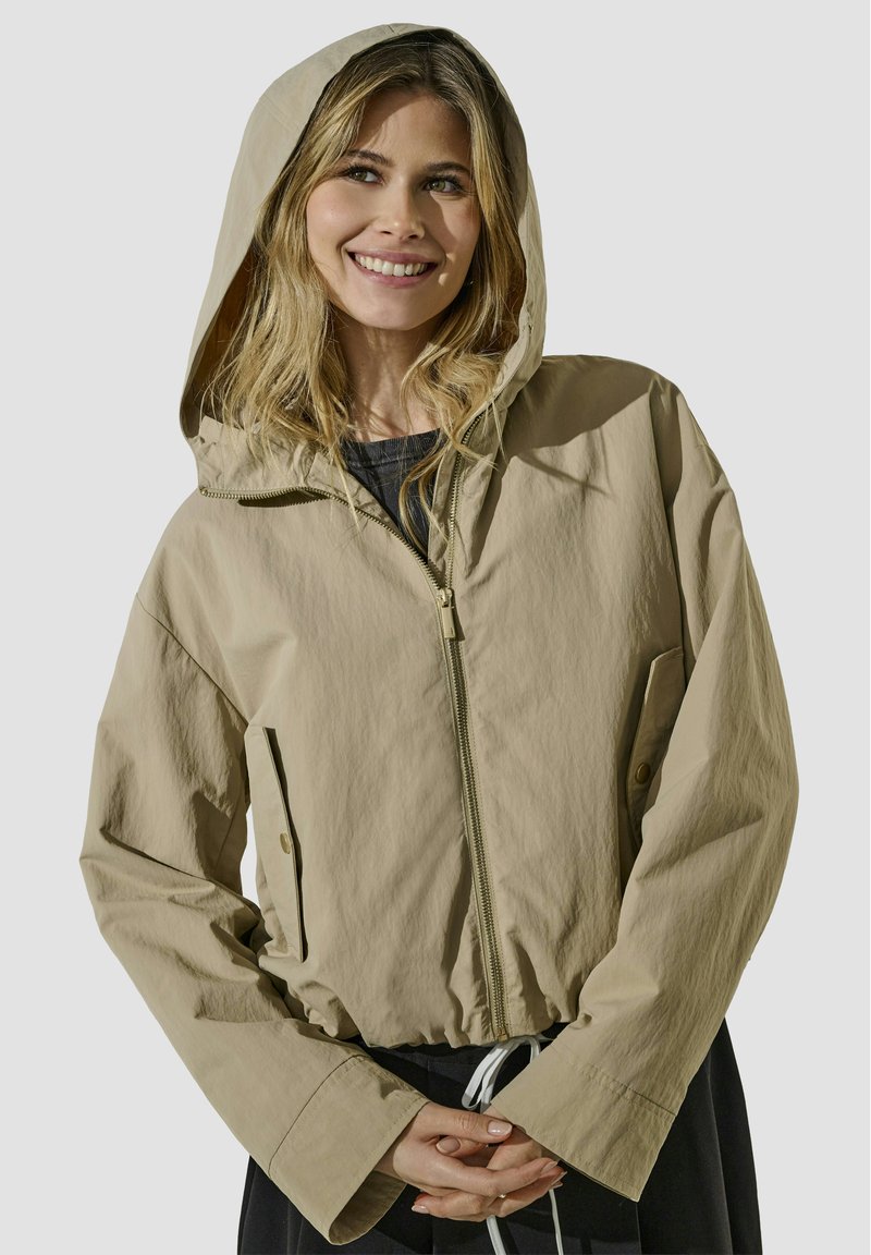 Veste légère beige avec fermeture éclair sur le devant, capuche et taille à cordon. Comprend des poches latérales et un tissu texturé.