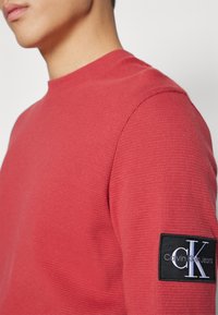 Röd stickad långärmad tröja med rund nacke och ribbat mönster. Har en svart rektangulär lapp med en vit logotyp på vänster ärm.