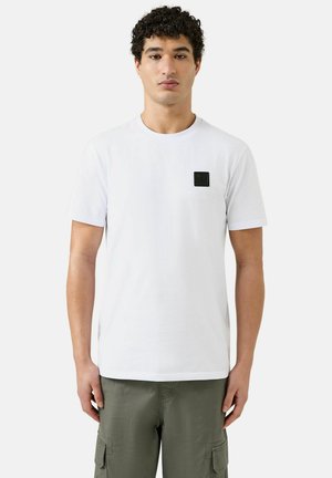 Jonge man met krullend haar, gekleed in een effen wit kortemouwen T-shirt en olijfgroene cargobroeken, staande tegen een witte achtergrond.