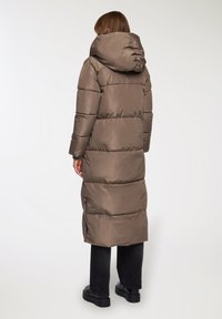 Lunga giacca puffer color taupe, con cappuccio, design trapuntato e cerniere ai lati. Indossata su pantaloni neri e scarpe nere chunky.