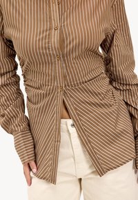 Brown and white striped button-up shirt with long sleeves, featuring ruched sides and a fitted design. Paired with cream-colored pants.