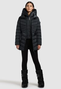 Sort puffersjakke med høj krave og hætte, med vandret quiltning, lynlås fortil og sidelommer. Bæres med sorte leggings og støvler.