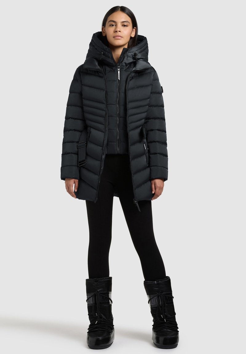 Sort puffersjakke med høj krave og hætte, med vandret quiltning, lynlås fortil og sidelommer. Bæres med sorte leggings og støvler.