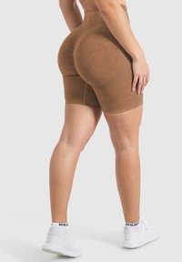 Pantaloni corti marroni a vita alta con una finitura liscia e texture, caratterizzati da un design che modella il corpo e una fascia elastica in vita, abbinati a sneakers bianche.