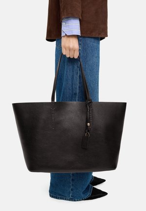 Czarna skórzana torba typu tote o wyraźnej strukturze, gładkiej fakturze, długich uchwytach oraz małej skórzanej zawieszce. Noszona z niebieskimi dżinsami.