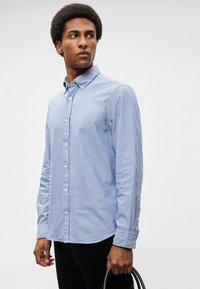 Ανοιχτό μπλε πουκάμισο με ρίγες, κατασκευασμένο από μαλακό ύφασμα, με κλασικό γιακά, μακρύ μανίκι και διακριτικούς κουμπιά που μοιάζουν με μαργαριτάρια.