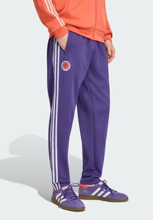 Osoba nosząca fioletowe spodnie dresowe z białymi paskami i czerwonobiałym emblematem, w połączeniu z fioletowymi butami Adidas i pomarańczową kurtką.