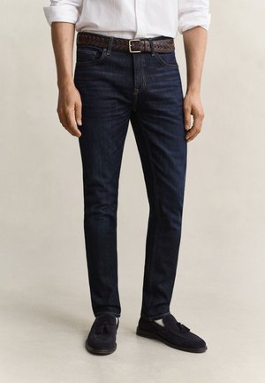 Homme portant un jean slim bleu foncé, une ceinture tressée marron, une chemise blanche aux manches retroussées et des mocassins noirs à pampilles, debout sur un sol clair.