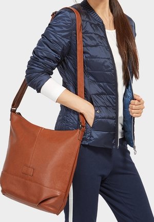 Vrouw draagt een navy blauw jasje en broek en draagt een grote bruine leren schoudertas met een lange riem over haar schouder.
