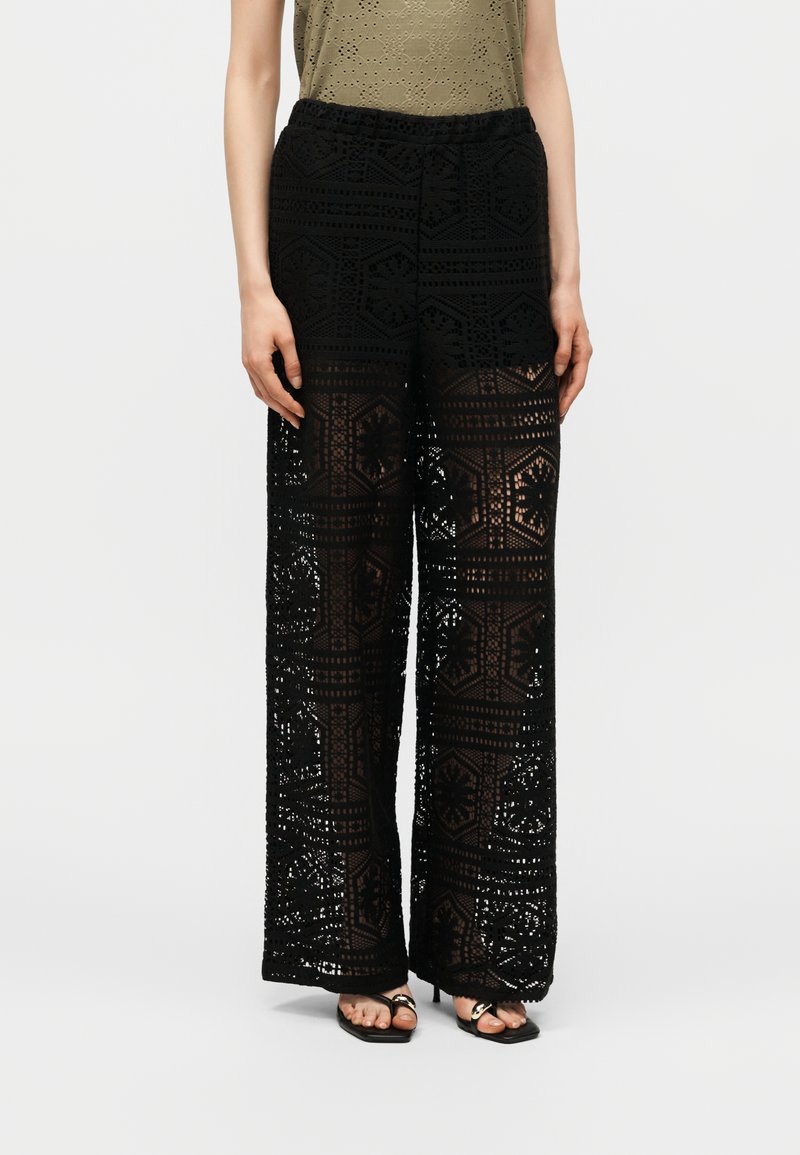 Femme portant un pantalon en dentelle noir à jambes larges avec des motifs géométriques et des talons ouverts noirs, associé à un haut olive à œillets.