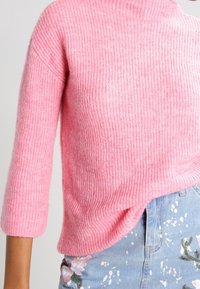 Rosa gestrickter Pullover mit gerippter Textur, dreiviertelÄrmel und asymmetrischem Saum, kombiniert mit hellen Blumenjeans.