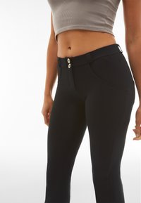 Nainen, jolla on yllään korkea vyötäröiset mustat housut, joissa on kaksoisnappikiinnitys, ja lyhyt, harmaa hihaton toppi valkoista taustaa vasten.