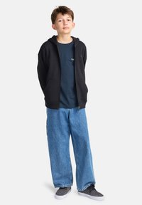 Felpa nera con zip sopra una maglietta blu scuro, abbinata a jeans denim chiari e larghi e scarpe da ginnastica nere. Outfit semplice e casual.