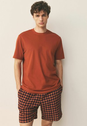 Młody mężczyzna z kręconymi włosami stoi przy gładkiej ścianie, ubrany w rdzawą koszulkę z krótkim rękawem i pomarańczowo-czarne szorty w kratę.