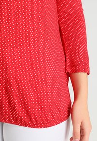 Rot-blaues Polka-Dot-Bluse mit glatter Textur, drei Viertelärmeln und abgerundetem Saum. Die Punkte sind weiß und gleichmäßig verteilt.
