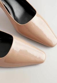 Beige lakota ādas augstpapēžu kurpes ar norādītu pirkstu, glītu spīdumu un melnu iekšējo oderi. Raksturīga eleganta un minimāla dizaina estētika.