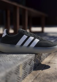 Scarpa da ginnastica nera con strisce bianche e suola traslucida che riposa su un gradino di metallo con texture in un ambiente esterno con sfondo sfocato.