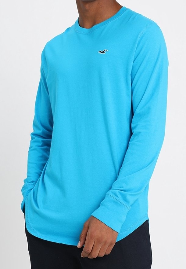 Long-sleeve t-shirt in bright turquoise fabric, featuring a rounded neckline and a small black logo on the chest. Smooth texture.