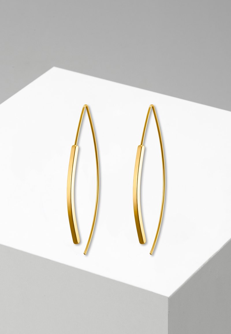 Paire de boucles d'oreilles fines et courbées en or avec un design minimaliste et allongé, présentée sur une surface géométrique blanche et grise.