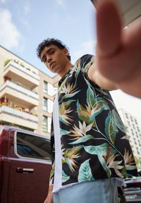 Young man in floral shirt reaching toward camera on urban street with buildings and vehicles in background.