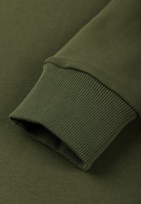 Lähikuva oliivinvihreän collegetakin hihan resorista, jossa on kohokuvioitu tekstiili ja taitettua sauman yksityiskohtaa.