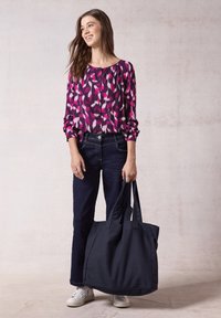 Blusa multicolore con motivi rosa e viola, jeans di denim scuro e una grande borsa nera con due manici. Scarpe da ginnastica bianche completano il look.