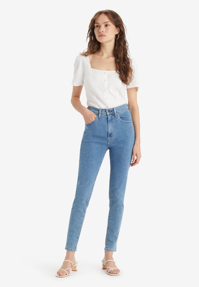 Jean en denim bleu taille haute avec une silhouette ajustée, comportant cinq poches et une fermeture à bouton. Associé à un haut blanc à manches courtes.