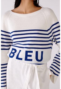 Pull en tricot blanc avec des rayures bleues et le texte « BLEU ». Il présente un col bateau et une coupe ample. Accentué par des manches larges à revers.