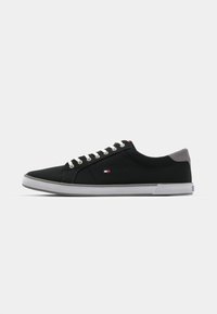 Zapatillas de lona negras con acentos grises, cordones blancos y un pequeño logo. Presentan una punta redondeada y una suela de goma en contraste.